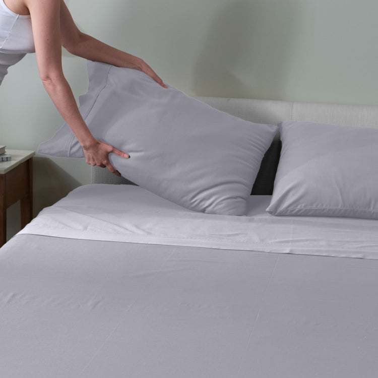A person is placing a light gray pillowcase on a neatly made bed with light grey sheets, shown in a bedroom setting. "Light Grey"