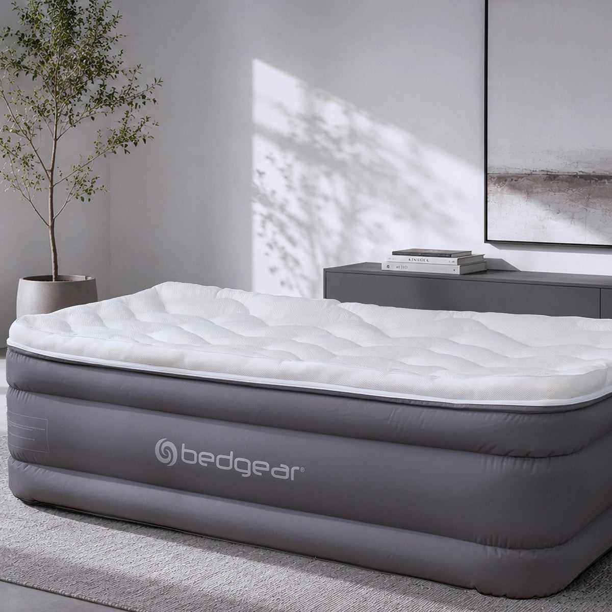 An air mattress by bedgear in dark gray, shown with a white quilted mattress topper in a modern bedroom setting with a sideboard, framed wall art, and a potted tree.