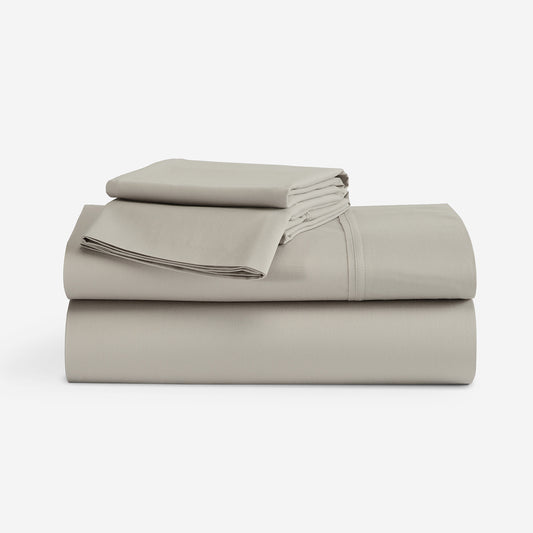 A stack of folded bedding sheets in a solid light taupe color, photographed on a seamless white studio background. "Taupe"