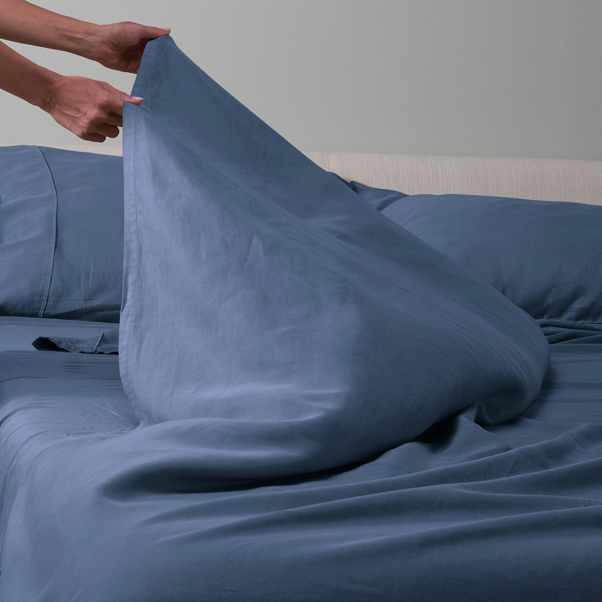 A person's hands lifting an indigo blue sheet over a matching blue bedsheet set on a bed with a light wood headboard.