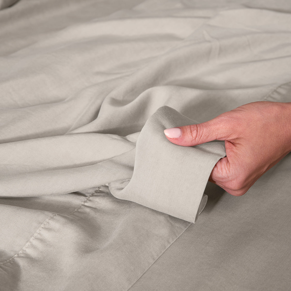 A close-up of a hand holding up a light beige bed sheet, showing the fabric texture and color.