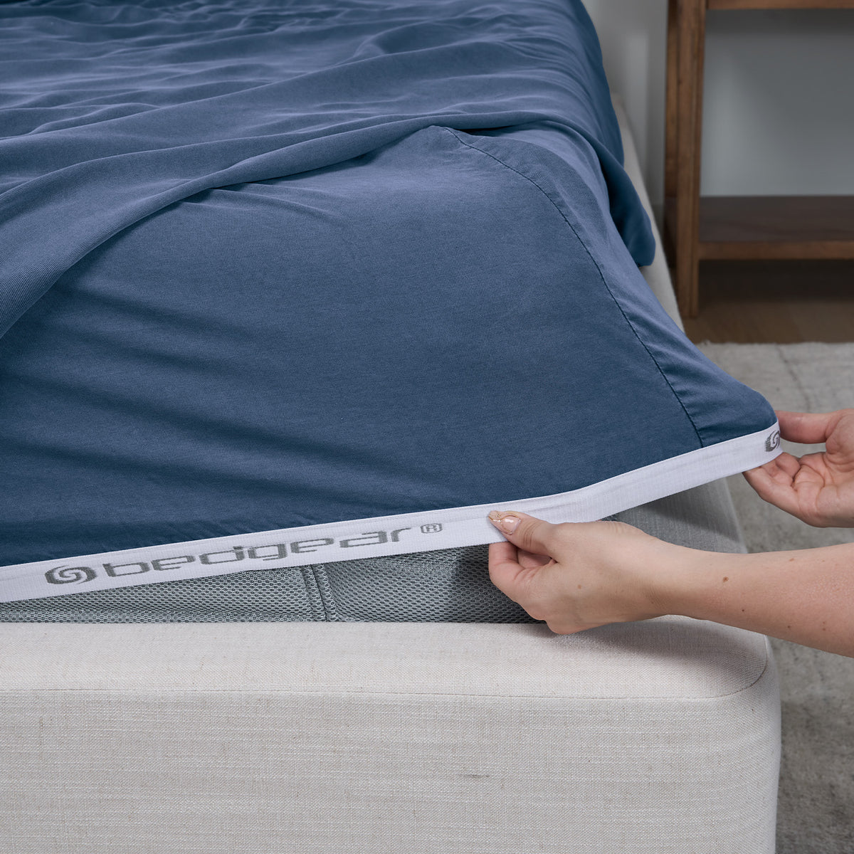A close-up of a bed with a fitted sheet being pulled up by hands. The sheet is an Indigo blue color with a white elastic band at the bottom edge that has the 'bedgear' brand name printed on it.