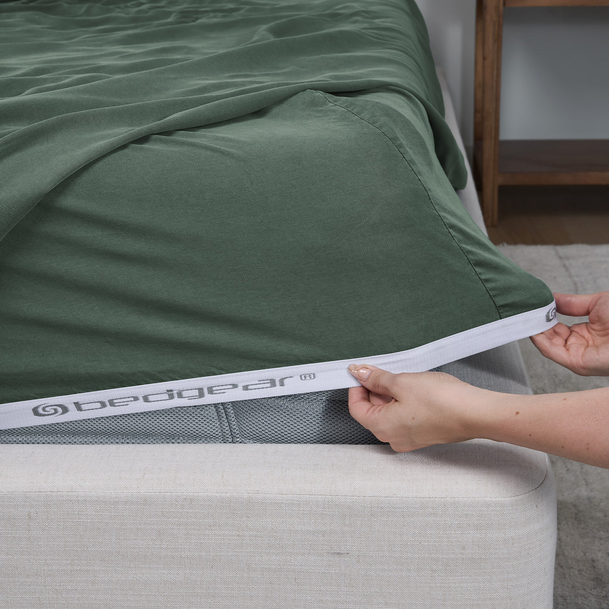 A close-up of a bed fitted with a moss green bed sheet, being pulled down over the mattress edge by hands. The sheet has a white elastic band with the brand name 'bedgear' printed on it.