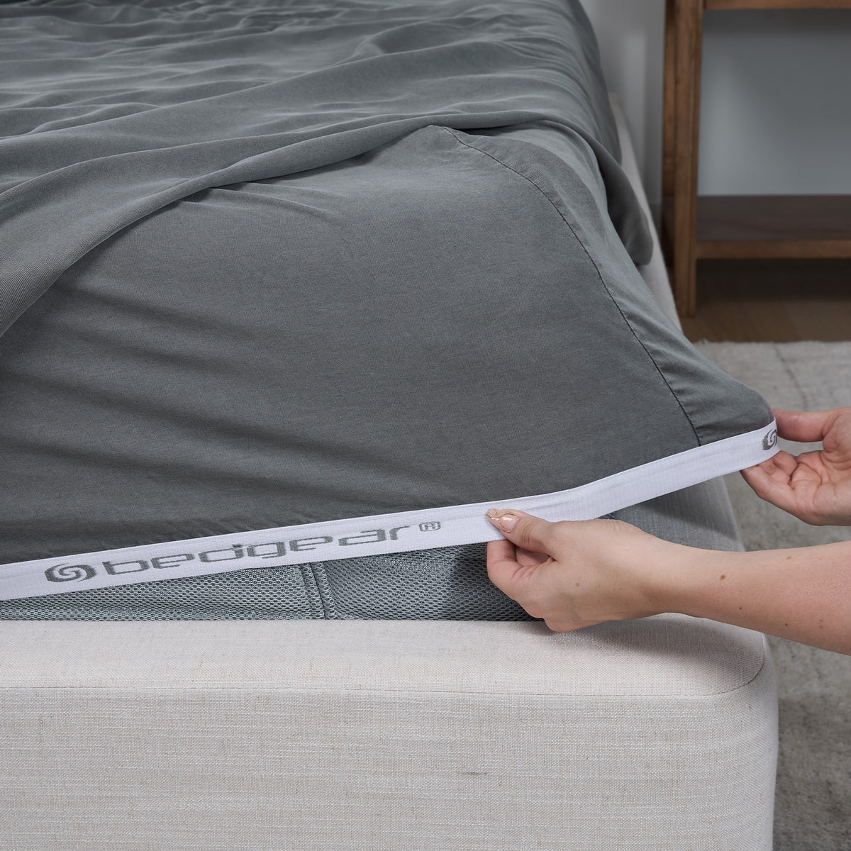 A close-up of a bed with a gray fitted sheet being pulled down over the mattress corner by hands, with the bed edge and a wooden bedside shelf visible in a bedroom setting.