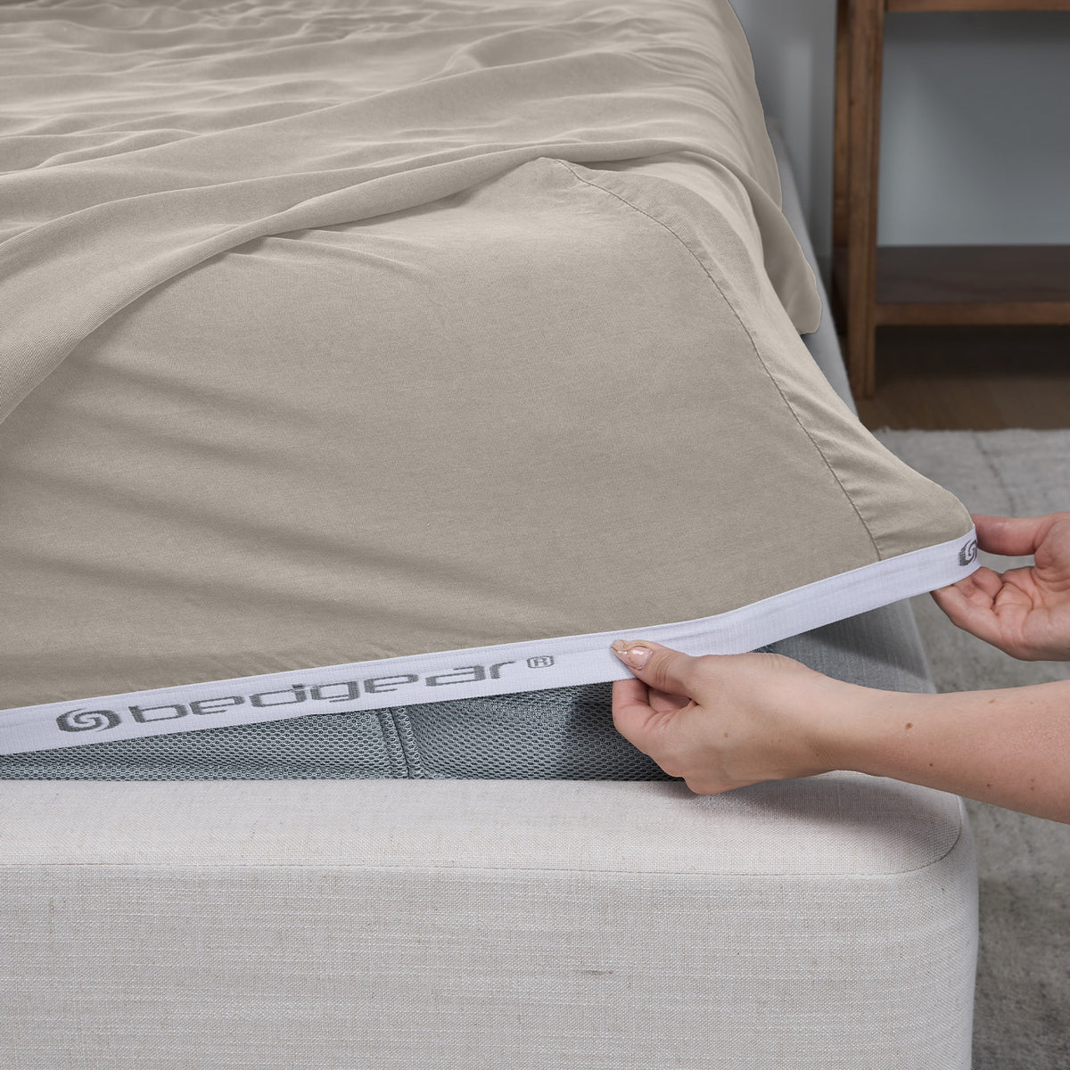 A close-up of a beige fitted sheet being pulled taut over the edge of a bed by hands, with the bedgear brand name visible on the white elastic band.