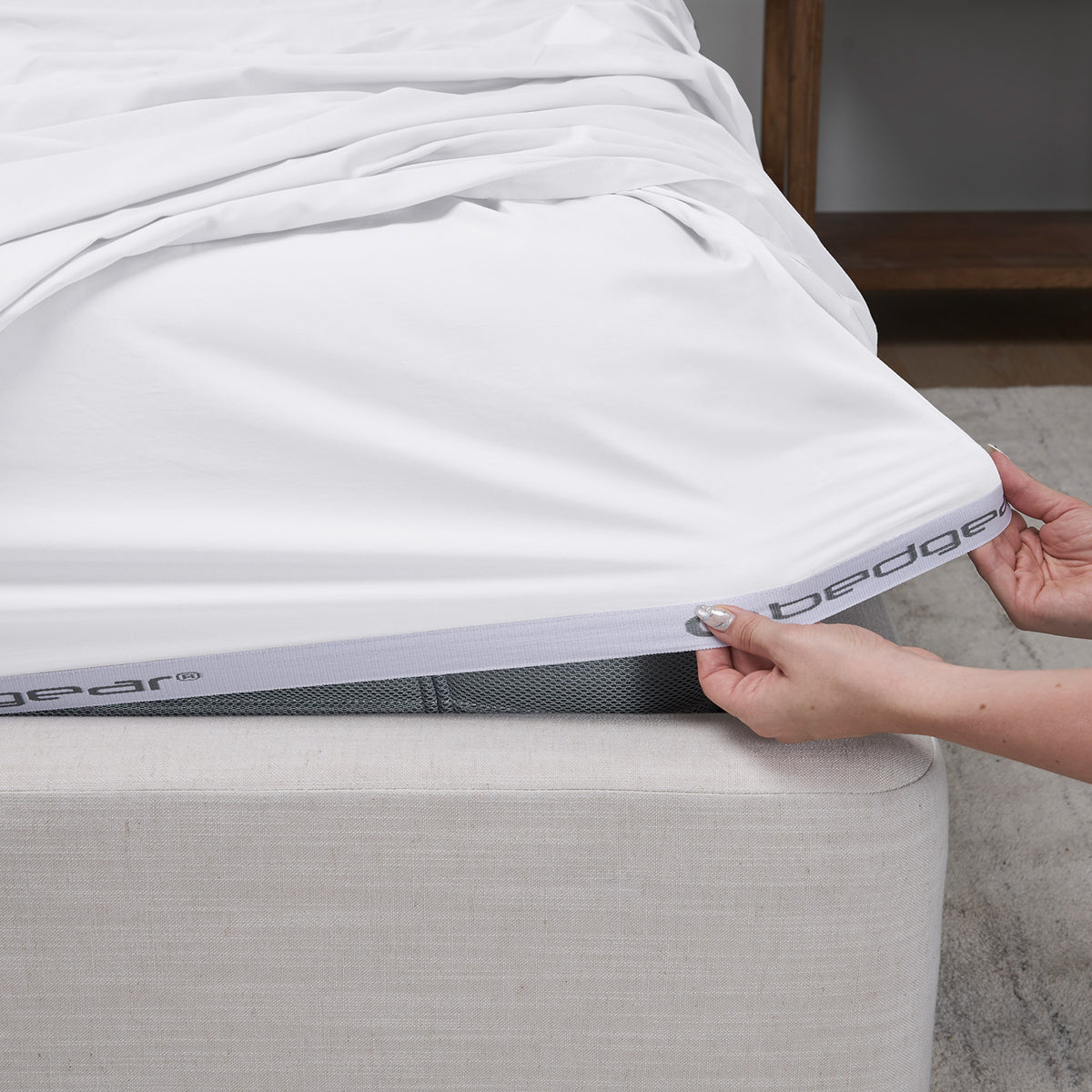A close-up of a white fitted sheet being pulled taut over a mattress corner by hands, with the bed frame and a neutral room background visible.