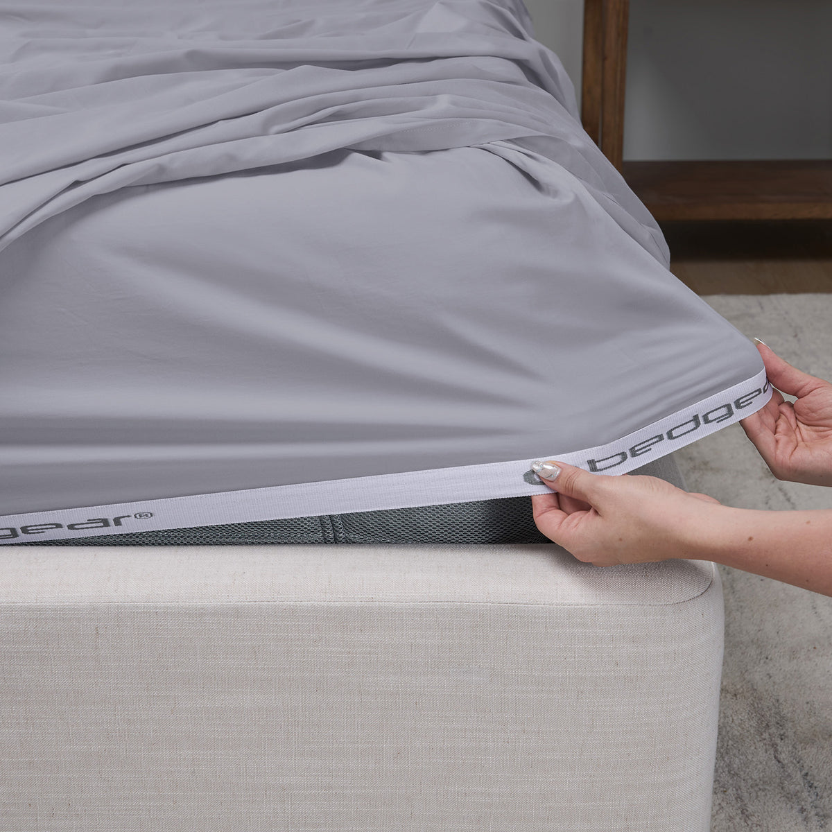 A close-up of a gray fitted sheet being pulled taut over the corner of a mattress by a person's hands. The sheet has a white elastic binding with Bedgear printed on it.