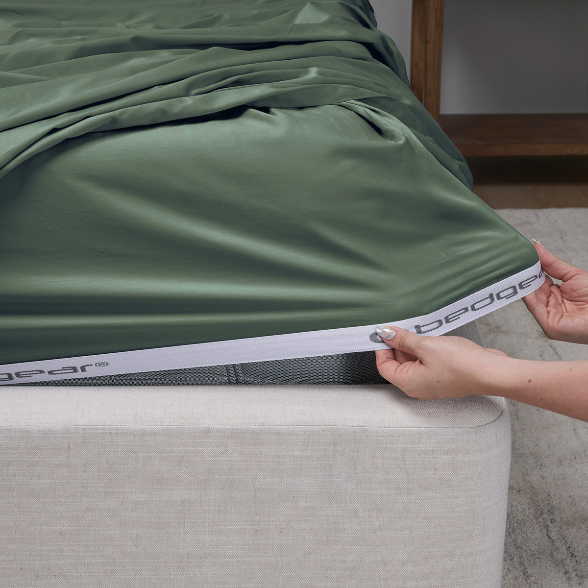 A moss green fitted sheet is being pulled taut over a mattress corner by a person's hands, with the Bedgear brand name visible on the white elastic band.