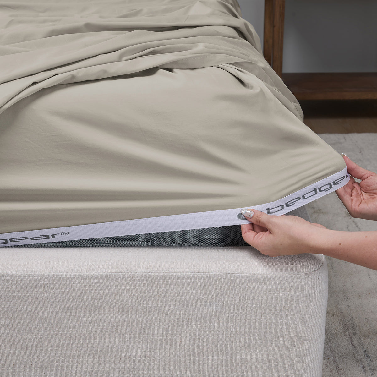 A close-up of a taupe  fitted sheet being pulled taut over the corner of a bed frame by a person's hands, with the Bedgear brand name visible on the elastic band.