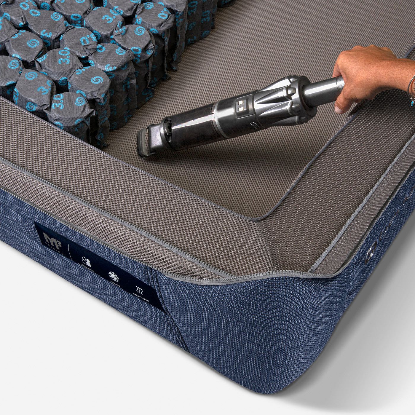 A close-up of a gray and blue mattress with a person lifting the corner, showing the interior structure and materials.