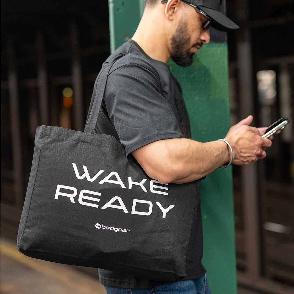 A man holding a Wake Ready tote bag and scrolling on his phone. 